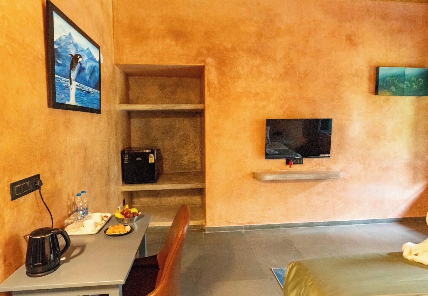 Una habitación de hotel con una cama, una mesa con un hervidor, un bol de fruta, una botella de agua, un televisor y un cuadro en la pared.