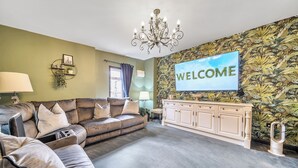 A living room with a sofa, a TV displaying 'WELCOME', a chandelier, and a patterned wall. - The Stables (Poulton-le-Fylde)
