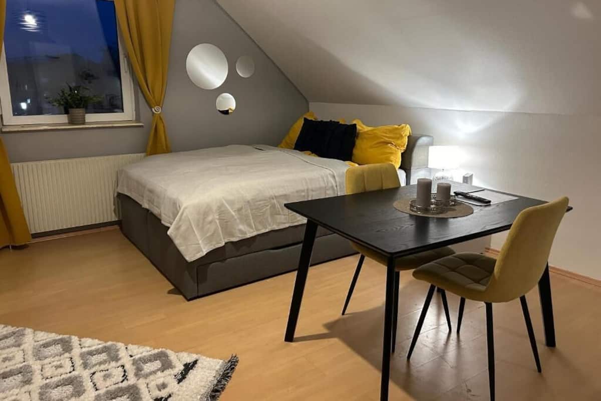Un dormitorio con cama, una mesa con silla, una ventana con cortinas y una lámpara fijada a la pared.