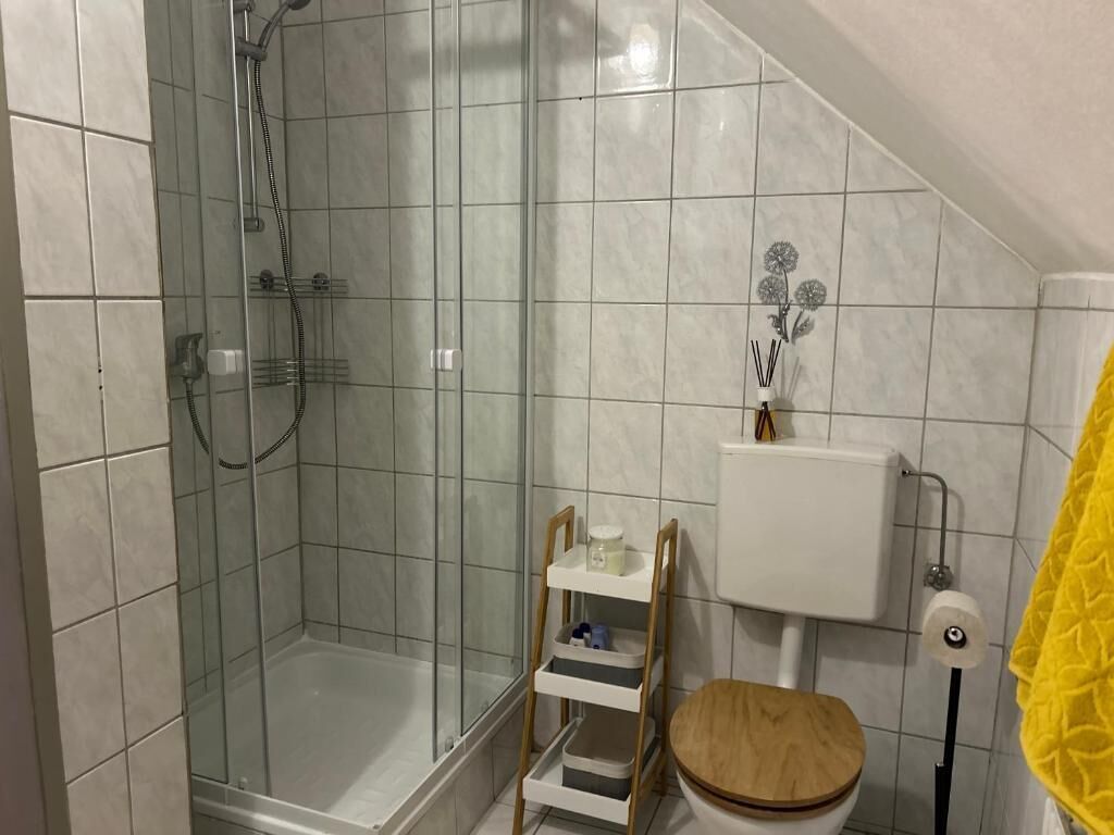 A bathroom with a glass-enclosed shower, a toilet with a wooden seat, and a white tiled wall.