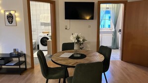 Uma área de refeições com uma mesa redonda, quatro cadeiras, um vaso com flores, uma TV de tela plana na parede e um relógio.