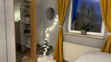Un dormitorio con cama, armario, espejo, ventana con cortinas y una pequeña planta en el alféizar.