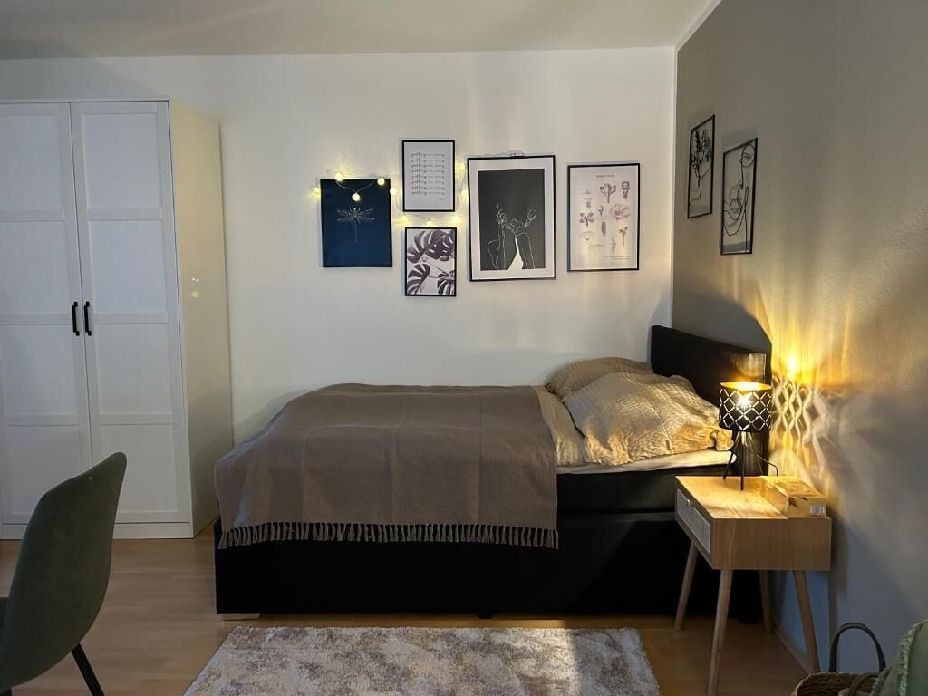 Een slaapkamer met een bed, nachtkastje, lamp, stoel en wanddecoratie.