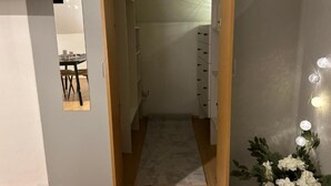 Um corredor com piso de madeira, parede branca e porta que dá para um quarto com cama e cômoda.