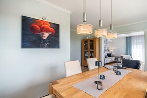 A dining area with a wooden table, white chairs, pendant lights, and a painting on the wall.