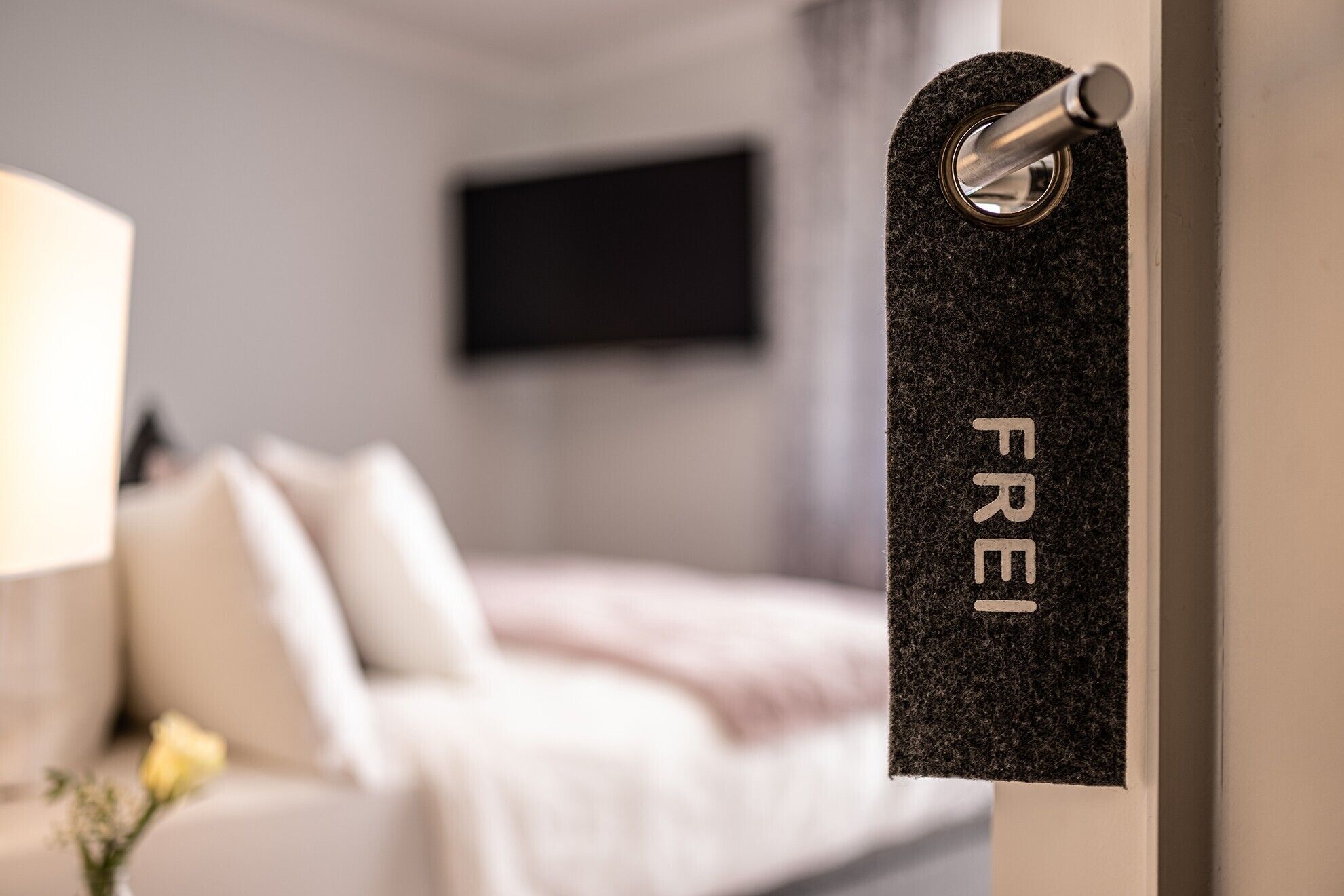 A hotel room with a bed, a TV mounted on the wall, and a door with a 'Frei' sign.