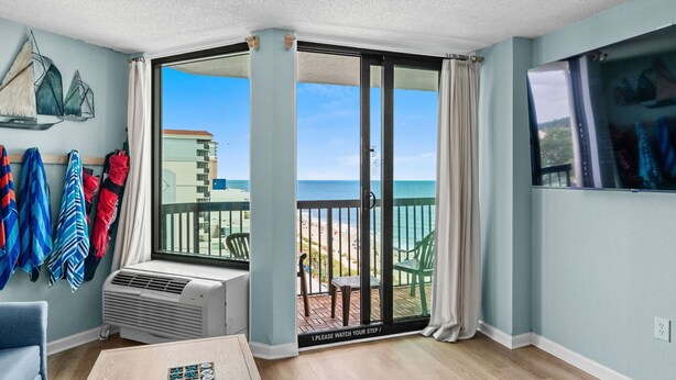 A room with a view of the beach, a sliding glass door, a table with a remote control, and a wall-mounted air conditioner. - Lighthouse Loft at Compass Cove - Unit 1151 (Myrtle Beach)