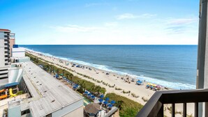 A beach view from a balcony with a clear sky, a sandy beach, and a few umbrellas. - Lighthouse Loft at Compass Cove - Unit 1151 (Myrtle Beach)