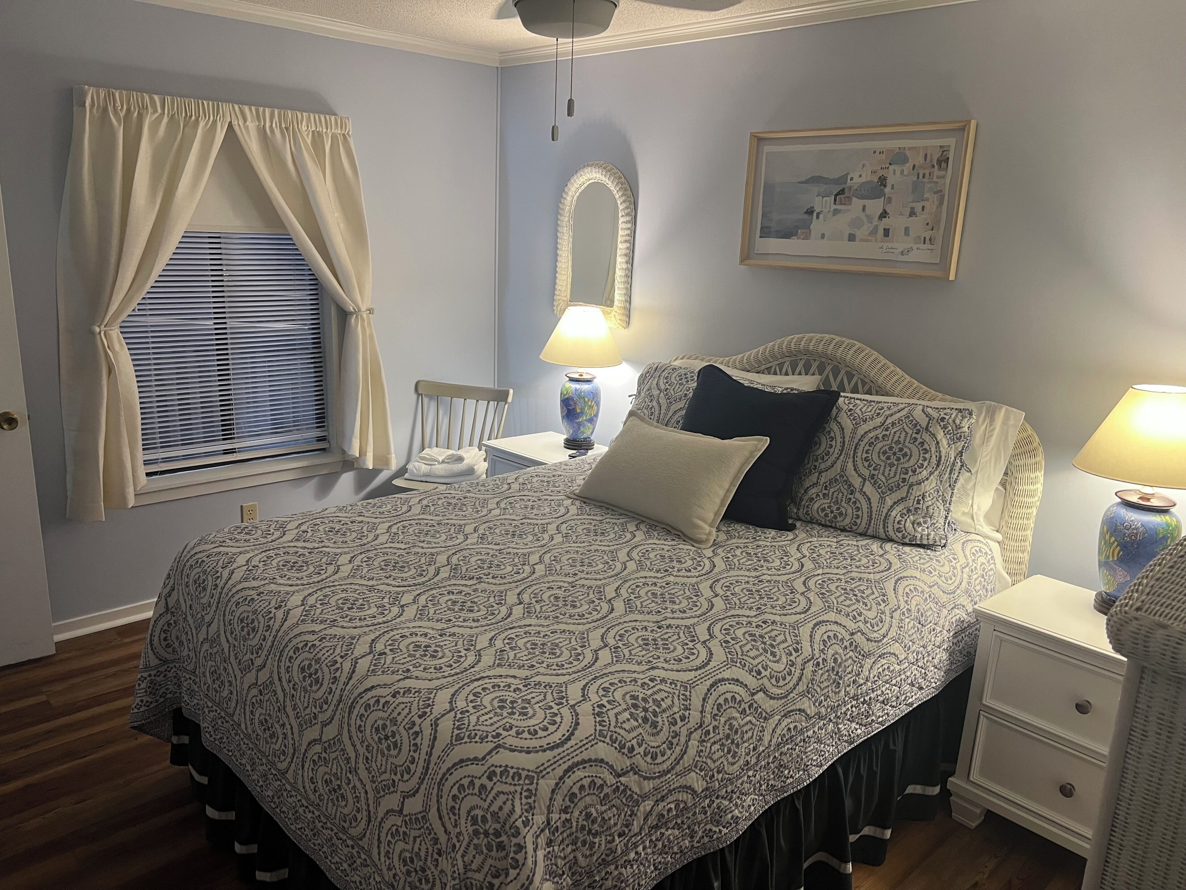 A bedroom with a patterned bedspread, a bedside lamp, a framed picture on the wall, and a window with curtains.