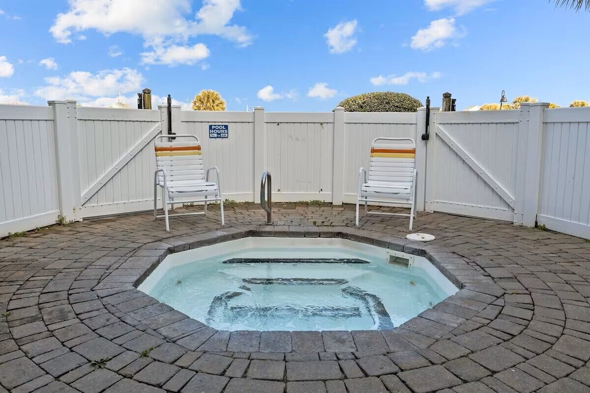 A hot tub with a brick patio, two white chairs, and a white fence with a pool hours sign.