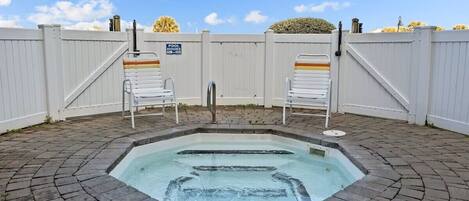 A hot tub with a brick patio, two white chairs, and a white fence with a pool hours sign.