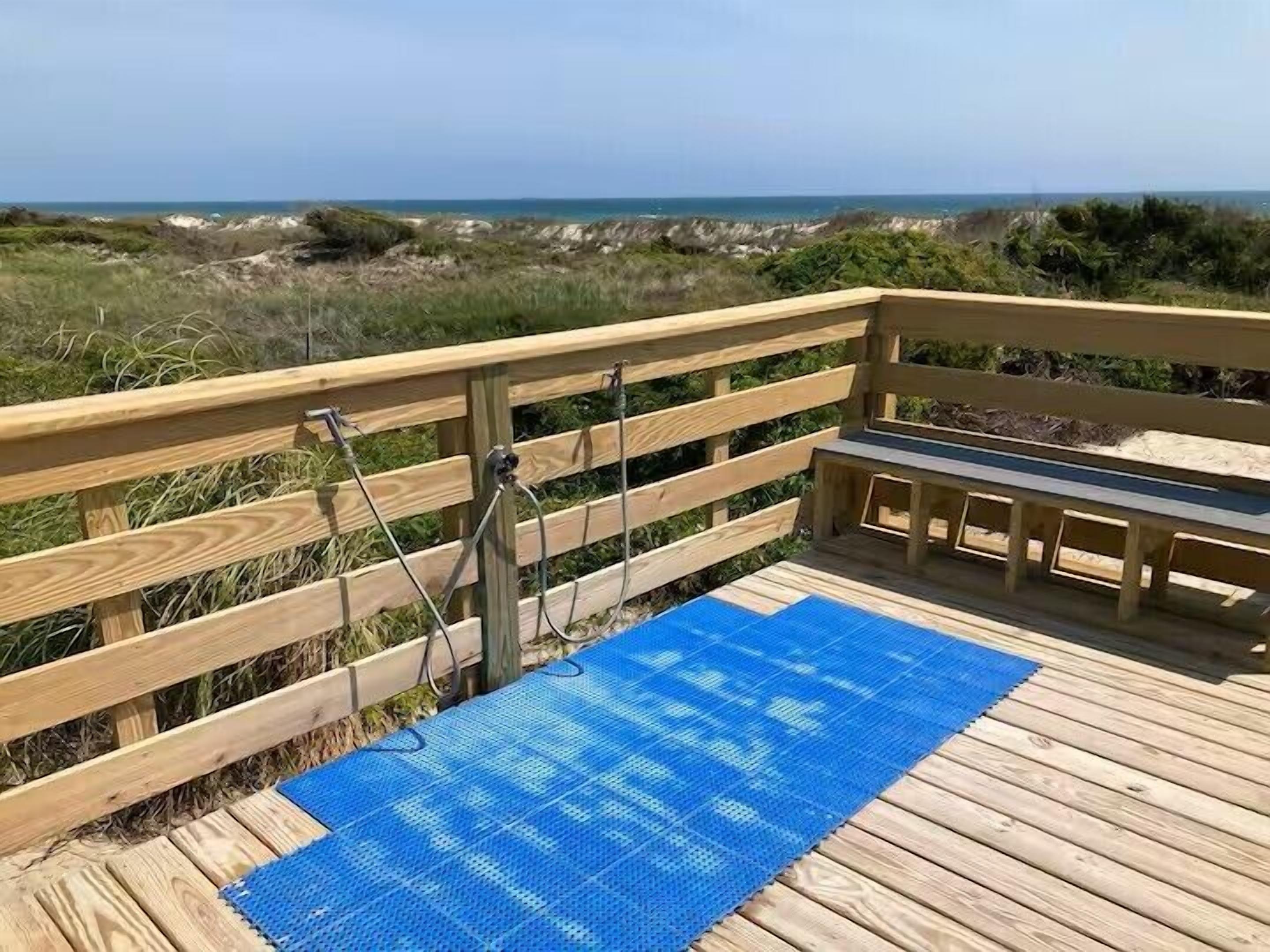 Eine Holzterrasse mit einer blauen Matte, einer Holzbank und einer Metallkonstruktion mit Blick auf Strand und Dünen.