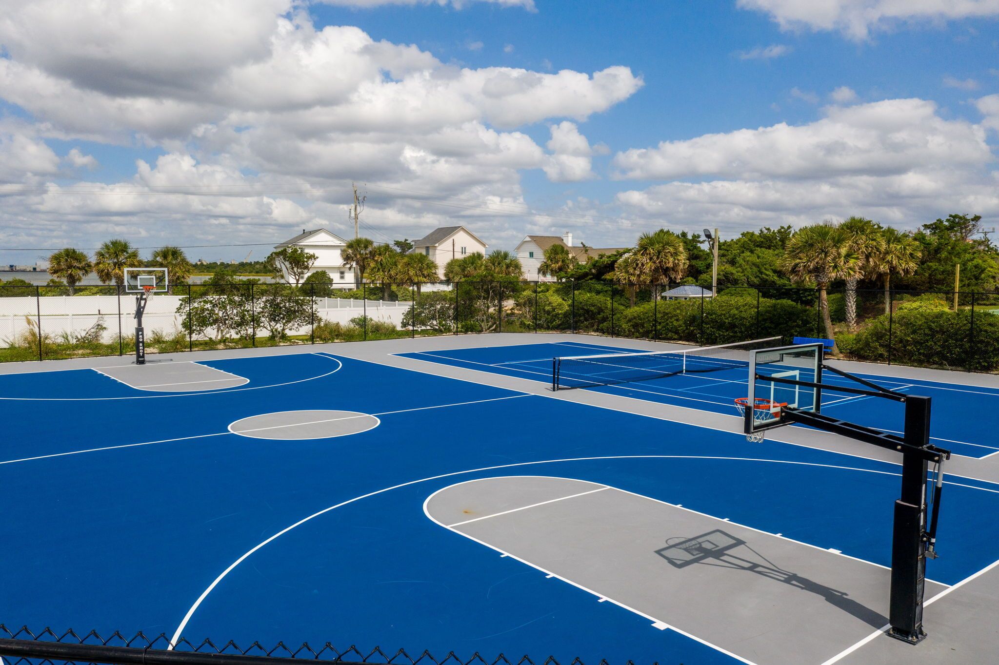 An outdoor basketball court with a blue surface, surrounded by a fence, and houses in the background.