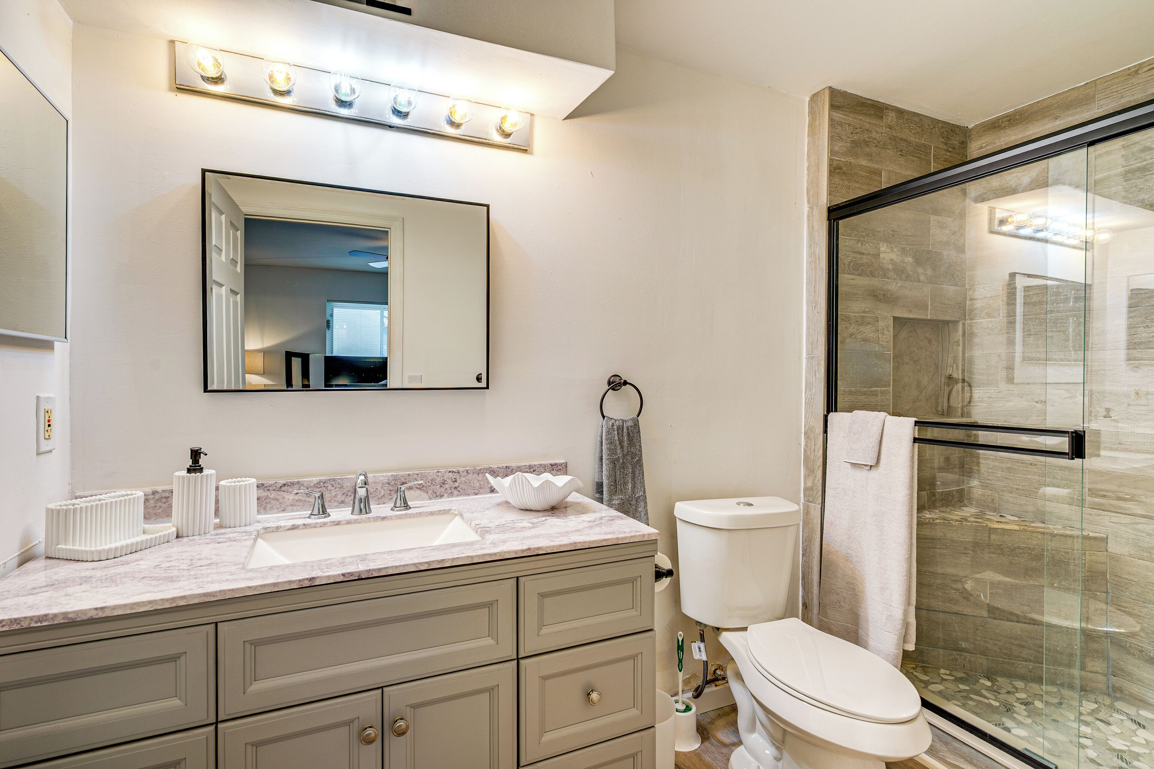 Une salle de bain dotée d’un plan de toilette en marbre, d’un lavabo, de toilettes, d’une cabine de douche en verre et d’un miroir avec des lumières au-dessus.