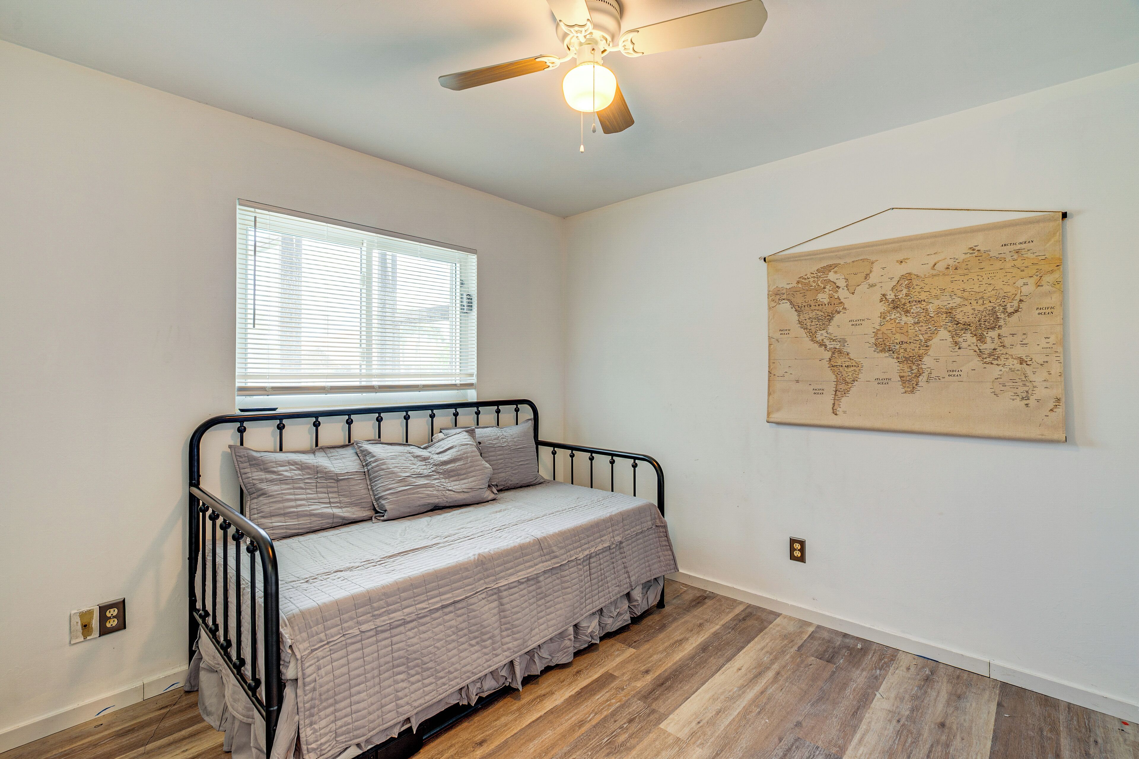 Une chambre avec un lit simple, un ventilateur de plafond, une fenêtre avec des stores et une carte du monde accrochée au mur.