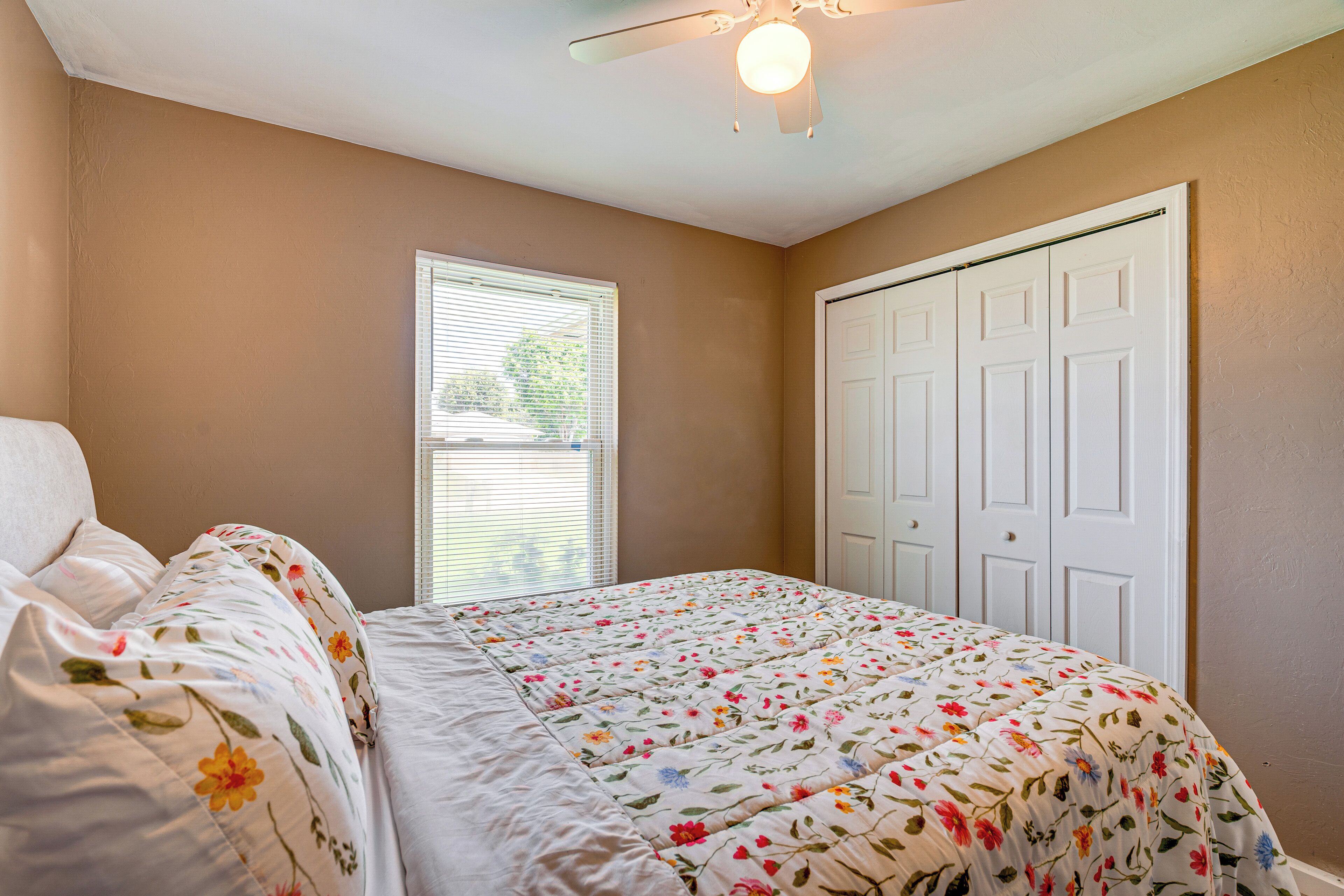 Ein Schlafzimmer mit Blumenmuster-Bettdecke, Deckenventilator, Fenster mit Jalousien und einer weißen Tür.