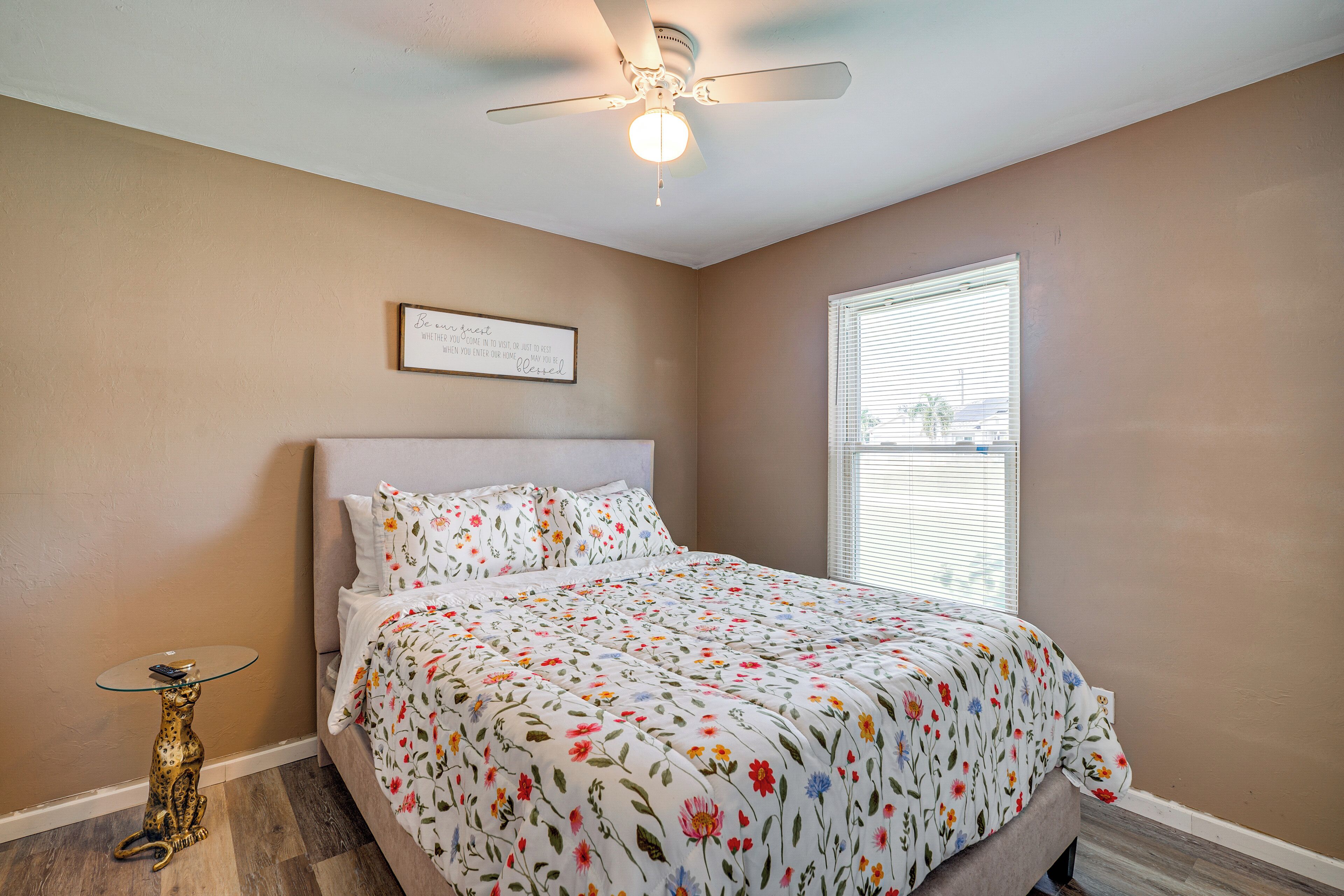 A bedroom with a bed, a ceiling fan, a window with blinds, and a small table with a decorative item.