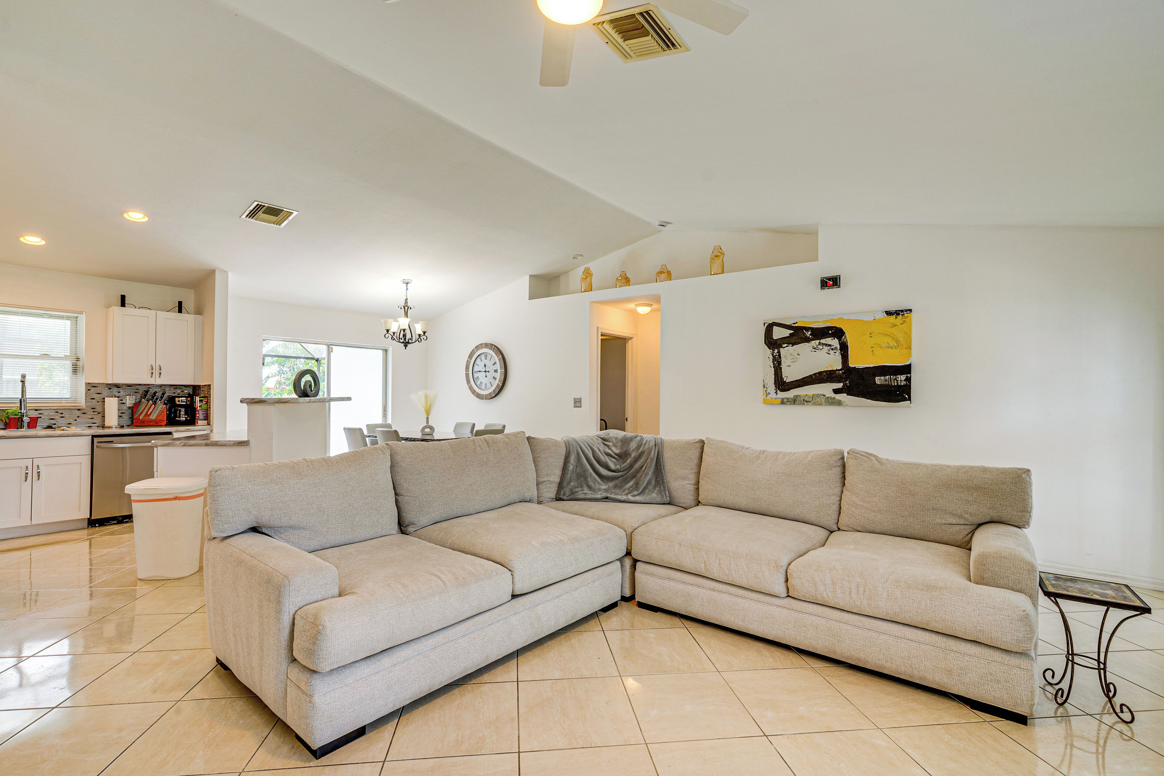 A modern living room with a beige sectional sofa, a dining area with a chandelier, and a kitchen with white cabinets.