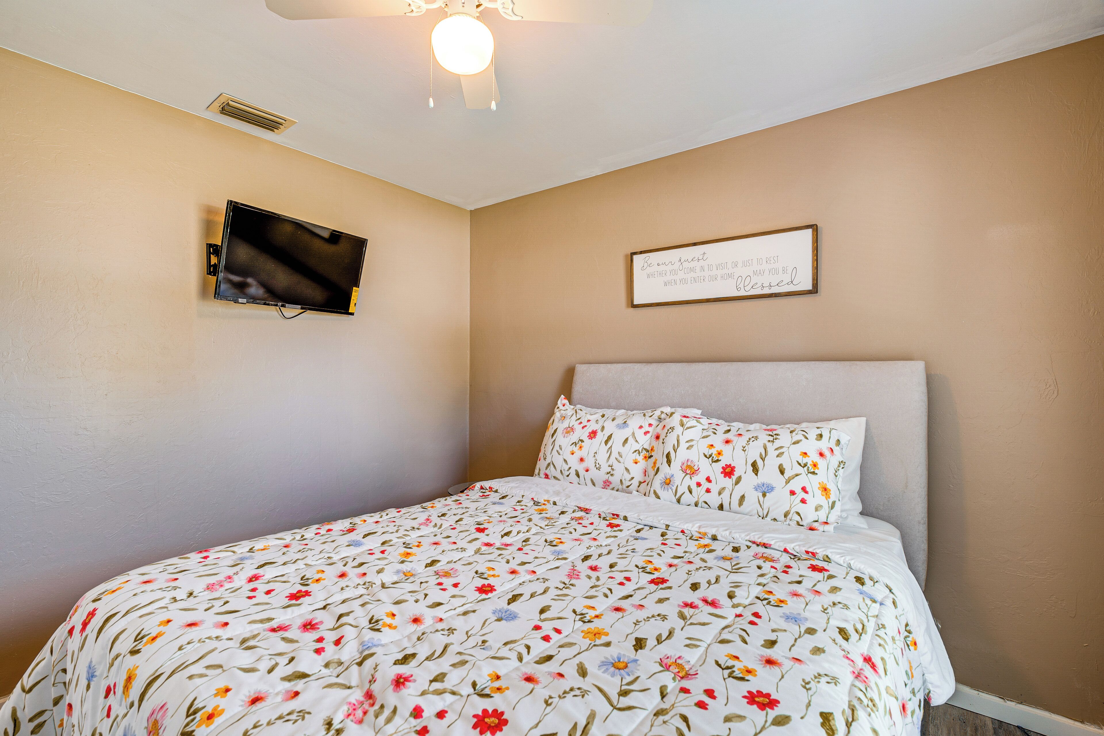 A bedroom with a bed, a wall-mounted TV, a ceiling light, and a framed sign.