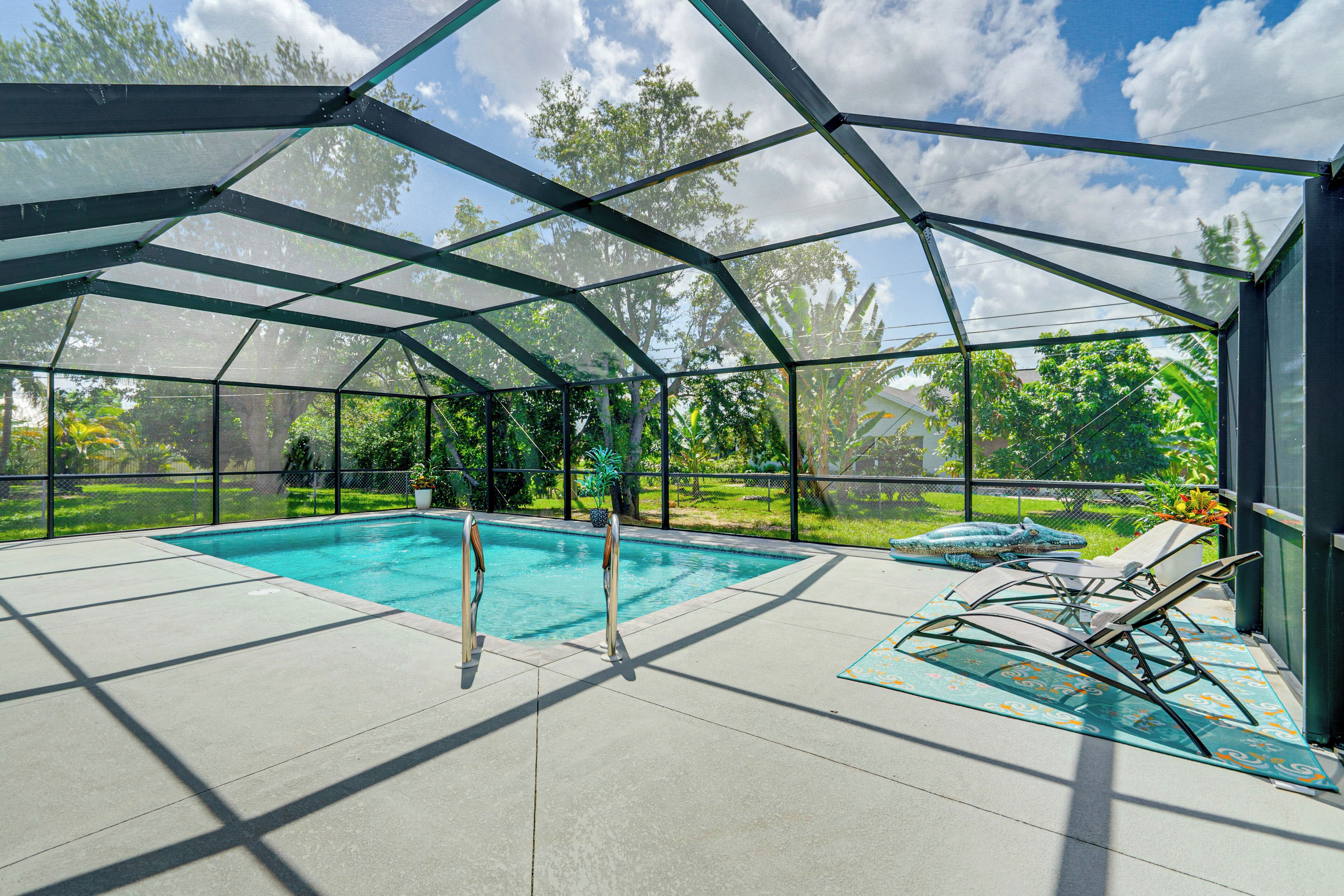 A covered outdoor swimming pool with a glass enclosure, surrounded by a patio area with lounge chairs and a view of a lush garden.