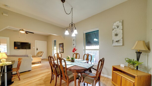 Un comedor con mesa de madera, sillas y una araña. Se ve un televisor al fondo y una ventana con vistas.