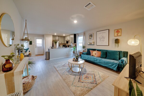 Una sala moderna con sofá, mesa de centro y televisor. Se ve una cocina al fondo con armarios y una ventana.