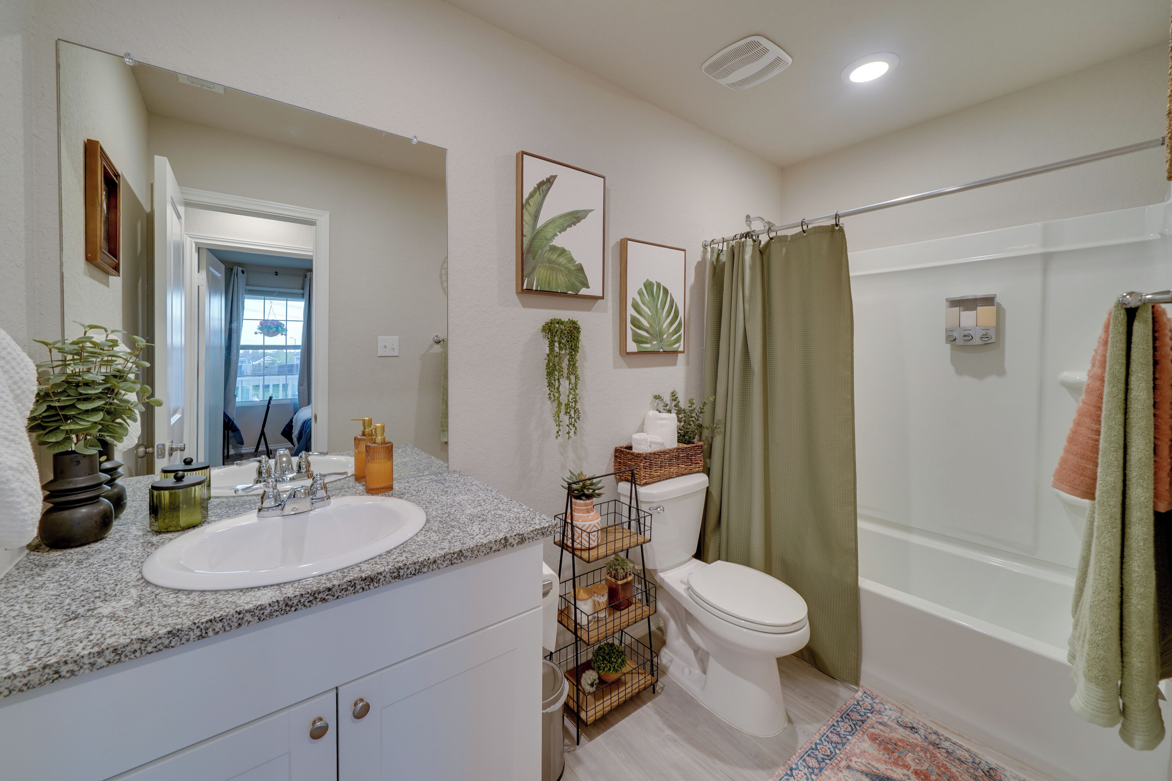 Une salle de bain avec un lavabo, des toilettes et une baignoire, tous de couleur blanche. On y trouve des plantes sur une étagère et un rideau de douche.