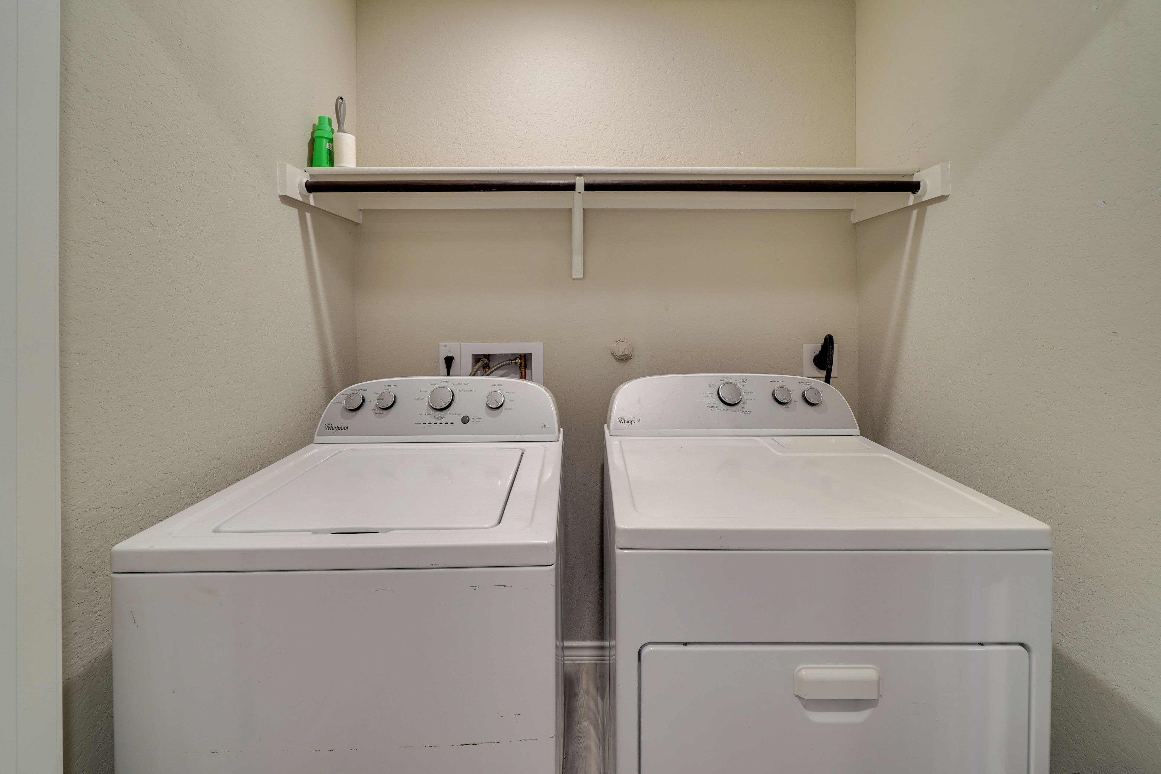 Pesuhuoneessa on kaksi valkoista Whirlpool-merkkiä olevaa pesu- ja kuivauskonetta, niiden yläpuolella hylly, jossa on vihreä pullo ja valkoinen astia, sekä valkoinen seinä.