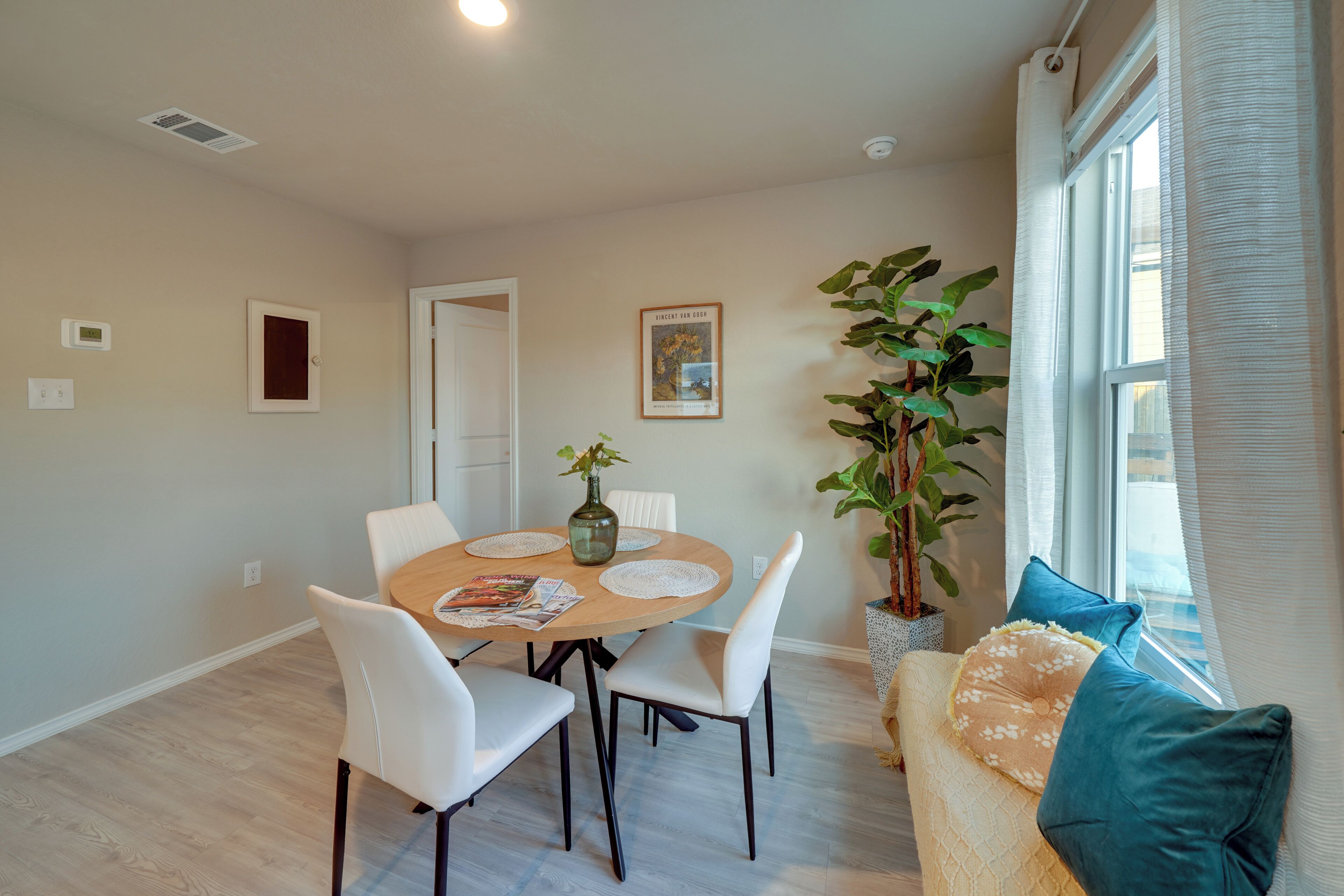A dining area with a round table, white chairs, a vase with a plant, and a window with curtains.