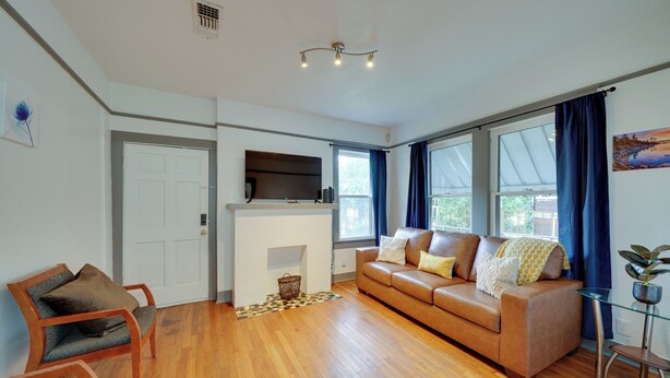 A living room with a brown leather sofa, wooden chairs, a flat-screen TV on a stand, a fireplace, and large windows with curtains. - 4 Mi to Downtown! Cozy Bungalow in San Antonio (San Antonio)