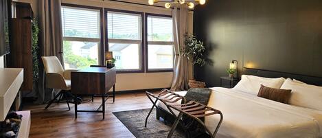 A modern bedroom with a large bed, a desk, and a chair. There is a window with blinds, a plant, and a ceiling light fixture.
