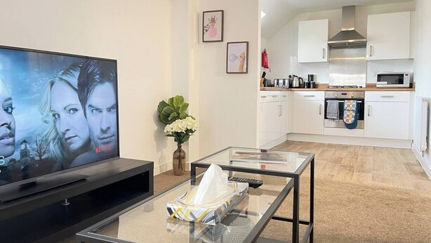 A modern living room with a flat-screen TV, a glass coffee table, and a kitchen area with white cabinets and appliances.