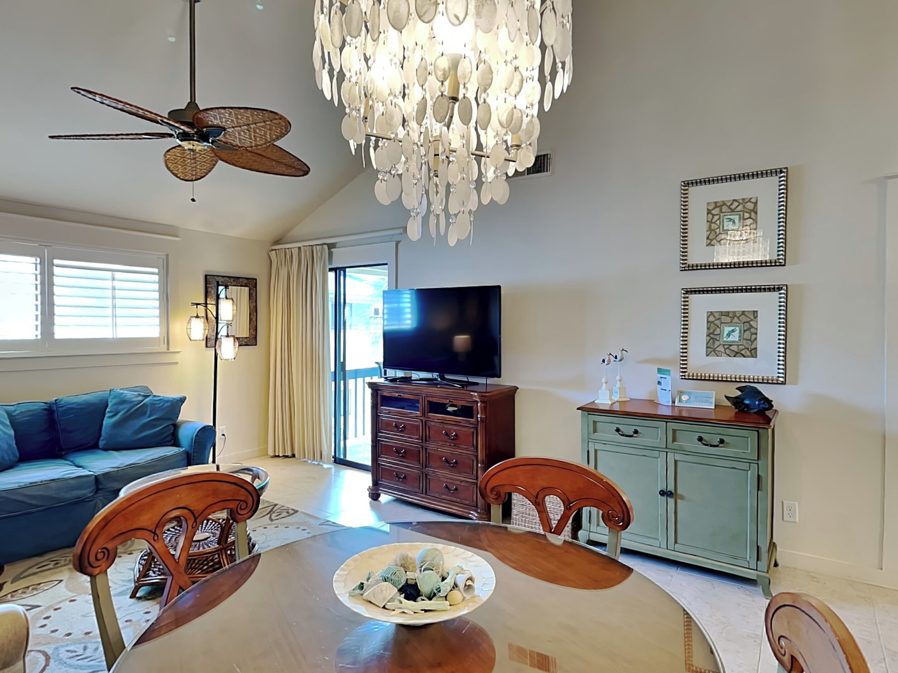 A spacious living room with a wooden dining table, a blue sofa, a ceiling fan, a chandelier, and a television.