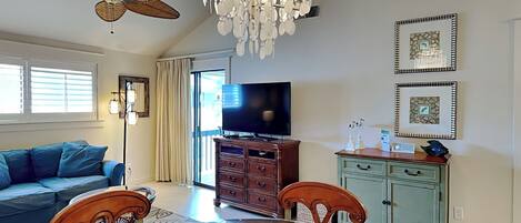 A spacious living room with a wooden dining table, a blue sofa, a ceiling fan, a chandelier, and a television.