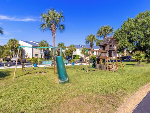A playground with swings and a slide, surrounded by palm trees and houses.