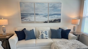 A living room with a white sofa, patterned throw, and a large ocean view painting. - Stylish Shadybrook retreat with golf cart, bikes & Tesla charger! (Sumterville)