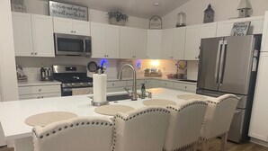 A modern kitchen with white cabinets, a stainless steel refrigerator, a microwave, and a sink. There are four bar stools with decorative stitching. - Stylish Shadybrook retreat with golf cart, bikes & Tesla charger! (Sumterville)