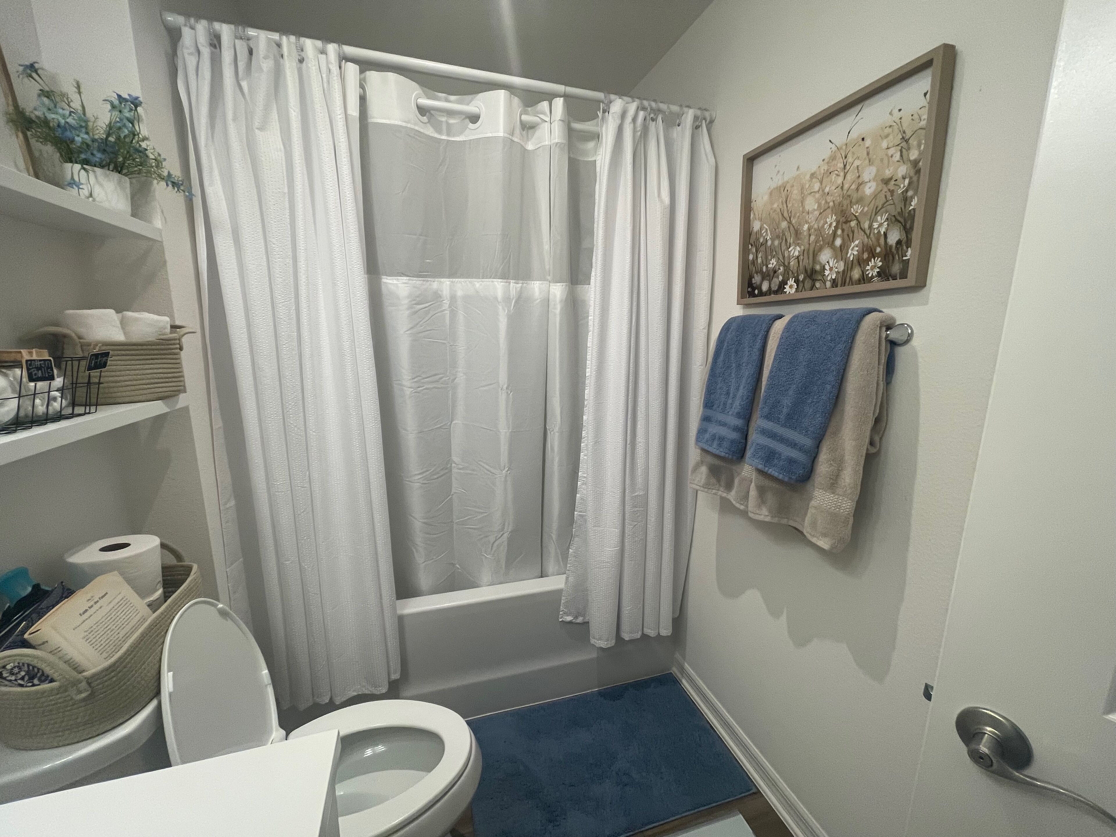 Ein Badezimmer mit Badewanne, Toilette und Waschbecken. Handtücher hängen an der Wand, und über der Badewanne hängt ein gerahmtes Bild.