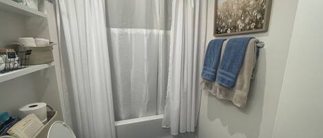 Ein Badezimmer mit Badewanne, Toilette und Waschbecken. Handtücher hängen an der Wand, und über der Badewanne hängt ein gerahmtes Bild.