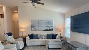 A living room with a white sofa, two armchairs, a coffee table, a patterned rug, a ceiling fan, wall-mounted artwork, and a TV on the wall. - Stylish Shadybrook retreat with golf cart, bikes & Tesla charger! (Sumterville)