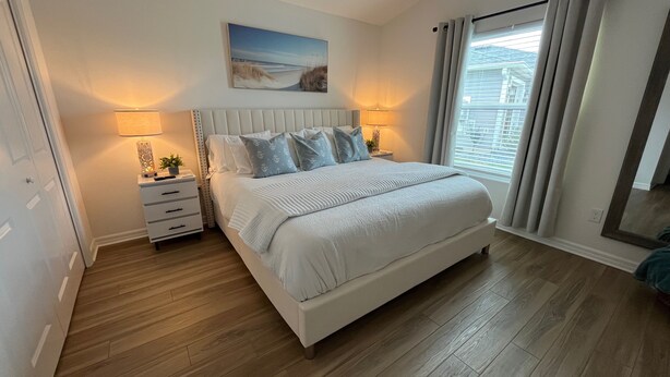 A bedroom with a large bed, bedside tables, a nightstand, a painting on the wall, and a window with curtains. - Stylish Shadybrook retreat with golf cart, bikes & Tesla charger! (Sumterville)