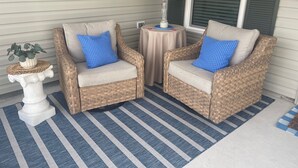 Two wicker chairs with blue cushions, a small white table with a lamp, and a striped rug on a porch. - Stylish Shadybrook retreat with golf cart, bikes & Tesla charger! (Sumterville)