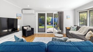 Een moderne woonkamer met een flatscreen televisie, een zwarte bank, een grijze bank en een blauwe slaapbank. Er is een schuifdeur die leidt naar een terras met uitzicht op een tuin.