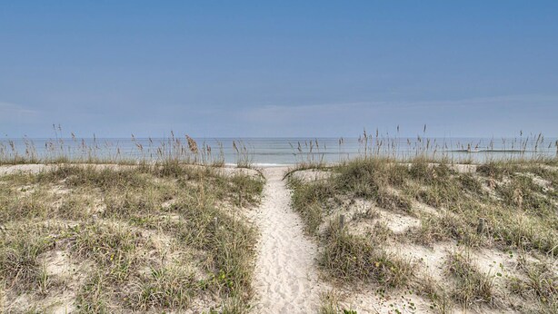 Uma trilha de areia que leva ao oceano, cercada por dunas baixas e vegetação rasteira.