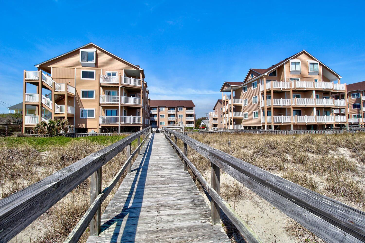A wooden boardwalk leading to a beachfront apartment complex with multiple balconies and a clear sky.