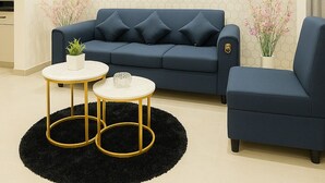 Uma sala de estar moderna com um sofá azul escuro, uma poltrona combinando e uma mesa de centro redonda com pés dourados. Um quadro grande de um leão com coroa está pendurado na parede.