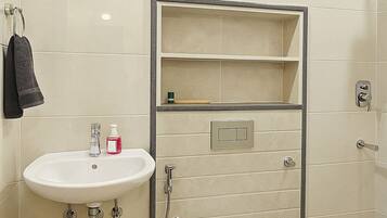 Une salle de bain avec un lavabo, des toilettes et une douche, tous de couleur blanche. On y trouve un porte-serviettes sur lequel est suspendue une serviette, ainsi qu’une étagère au-dessus du lavabo.