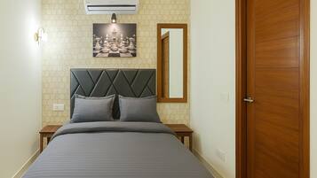 A bedroom with a bed, a headboard, a nightstand, a wall-mounted air conditioner, a wall-mounted light, a framed picture, a mirror, and a wooden door.