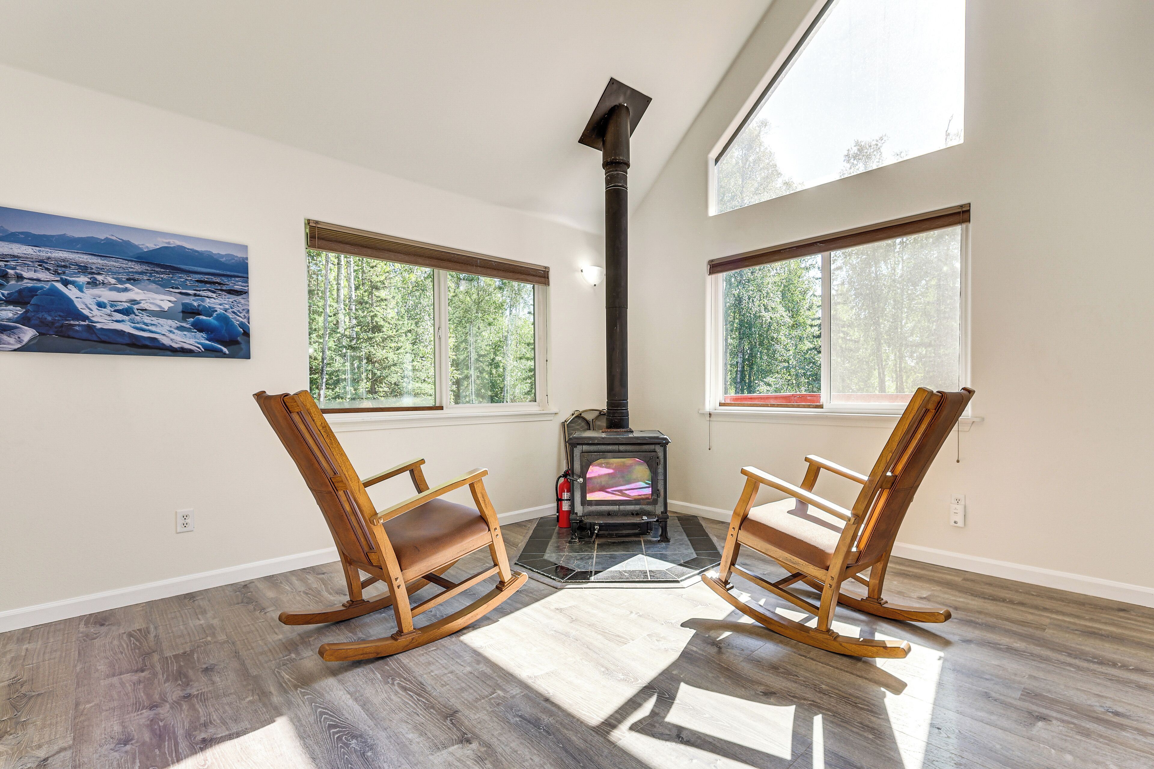 A room with a wood-burning stove, two rocking chairs, large windows, and a painting on the wall.