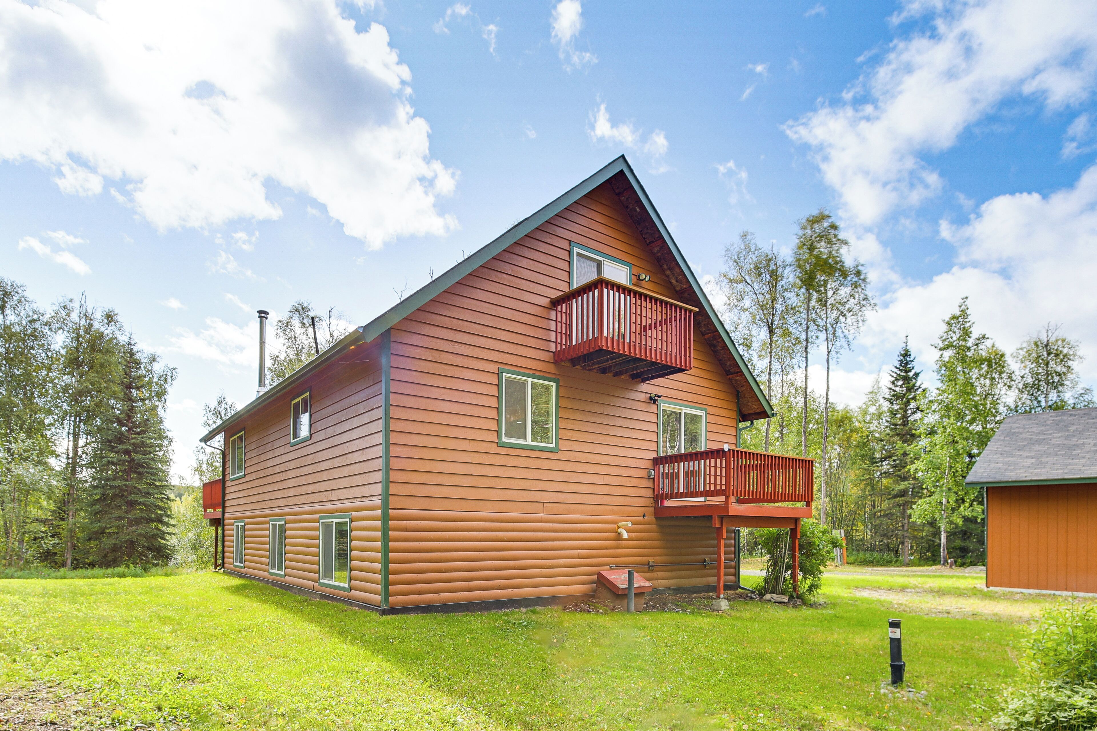 Une cabane en rondins à deux étages, dotée d’un balcon, entourée d’une cour verdoyante et d’arbres.
